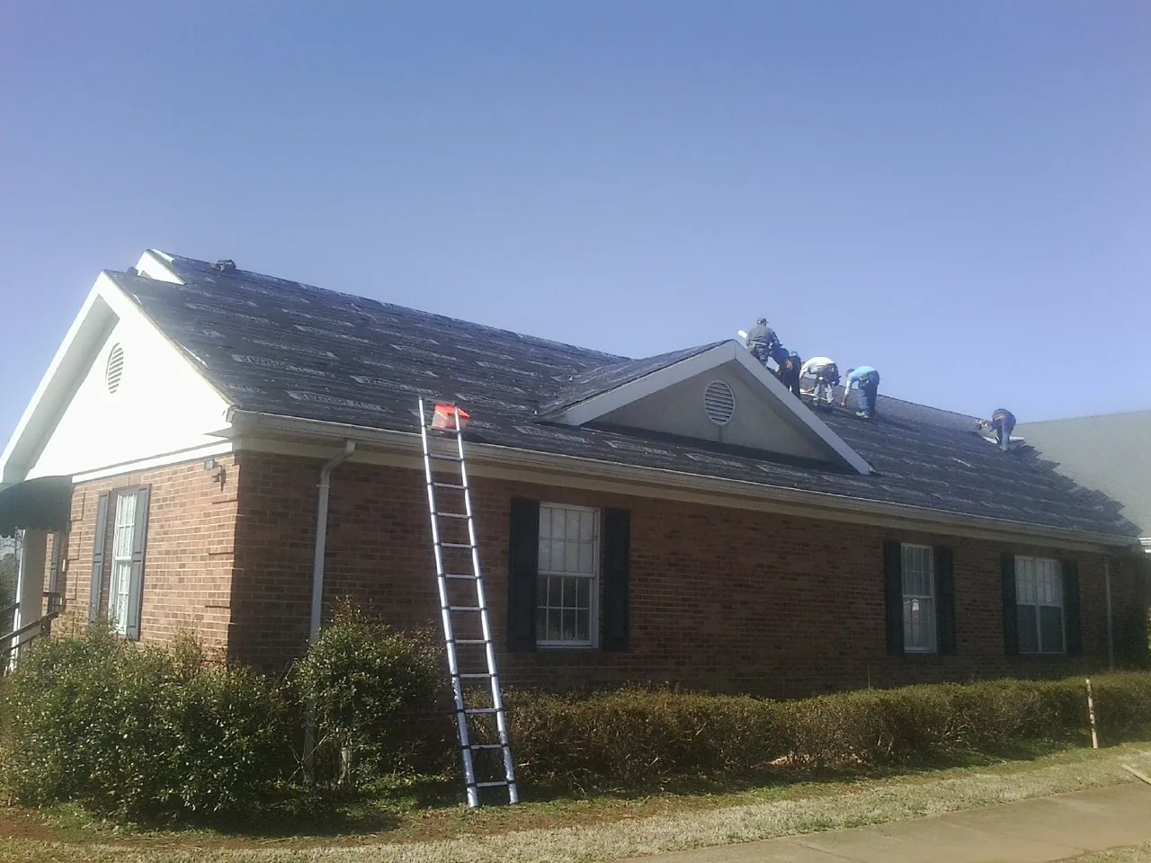 Crew tearing shingles off a brick ranch roof with a ladder set — Metal Roof Repair work by our crew in Simla