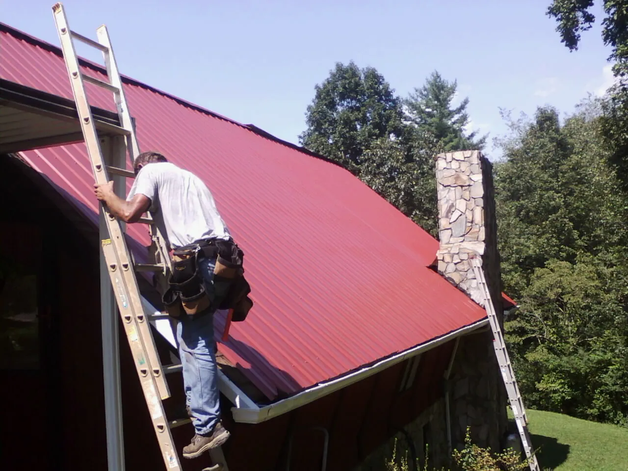 Roofer on a ladder beside a red metal roof and stone chimney — Soffit & Fascia Repair by Roofing Brothers in Simla